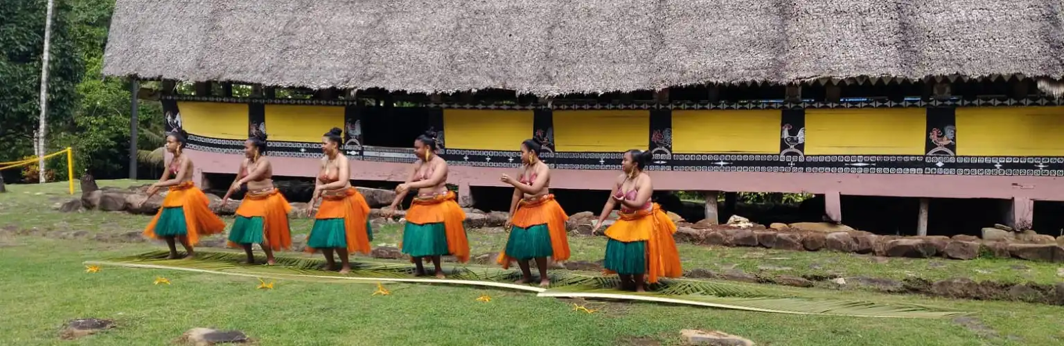 6 Female dancers in traditional grass skirts in front of a Palauan Men's House a so called Bai