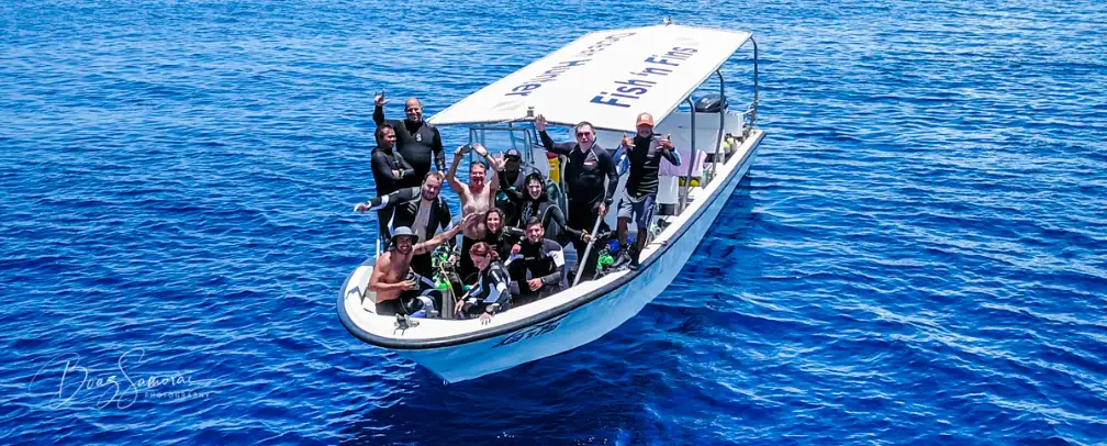 Scuba divers standing on deck of a Fish 'n Fins boat after a dive waving at the camera, which may be 20 meters away, the whole boat is visible from about 10 degrees front angle