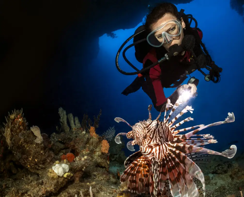 underwater photo of a female scuba diver with a torch, in the foreground a lion fish, the scene is in a cave